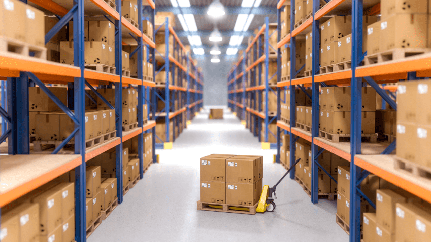 A warehouse aisle lined with pallet racking stacked with cartons, and a pallet truck carrying boxes in the middle of the aisle